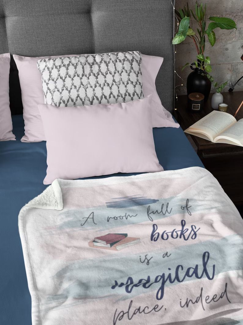 Photo of a blanket draped over a bed. The blanket is pink and blue with an illustration of a stack of books, and the text A room full of books is a magical place, indeed 