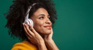 light brown-skinned Black woman with big fro holds manicured hands to white over-the-ear headphones while smiling; background is green