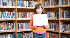 child in a library holding up a picture book standing in front of bookshelves