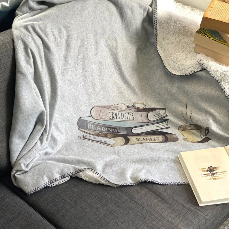 Photo of a grey blanket with white fuzzy interior. The print is a stack of 3 books with glasses on top and a coffee mug next to it, the stack reads on the spine of each book Granpa's Reading Blanket