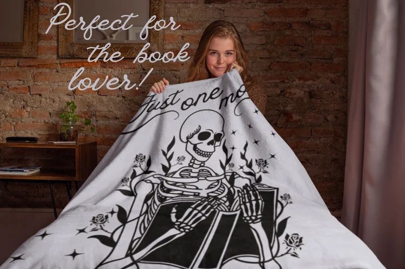Photo of a person holding a blanket showing the illustration of a skull reading a book, with the text Just one more chapter on top