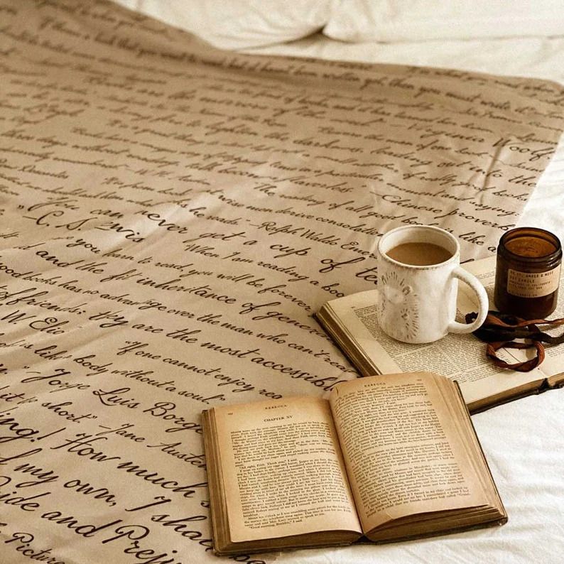 Photo of an open blanket with a couple of books, a mug and a jar placed on it. The blanket has several quotes in it, resembling an old letter