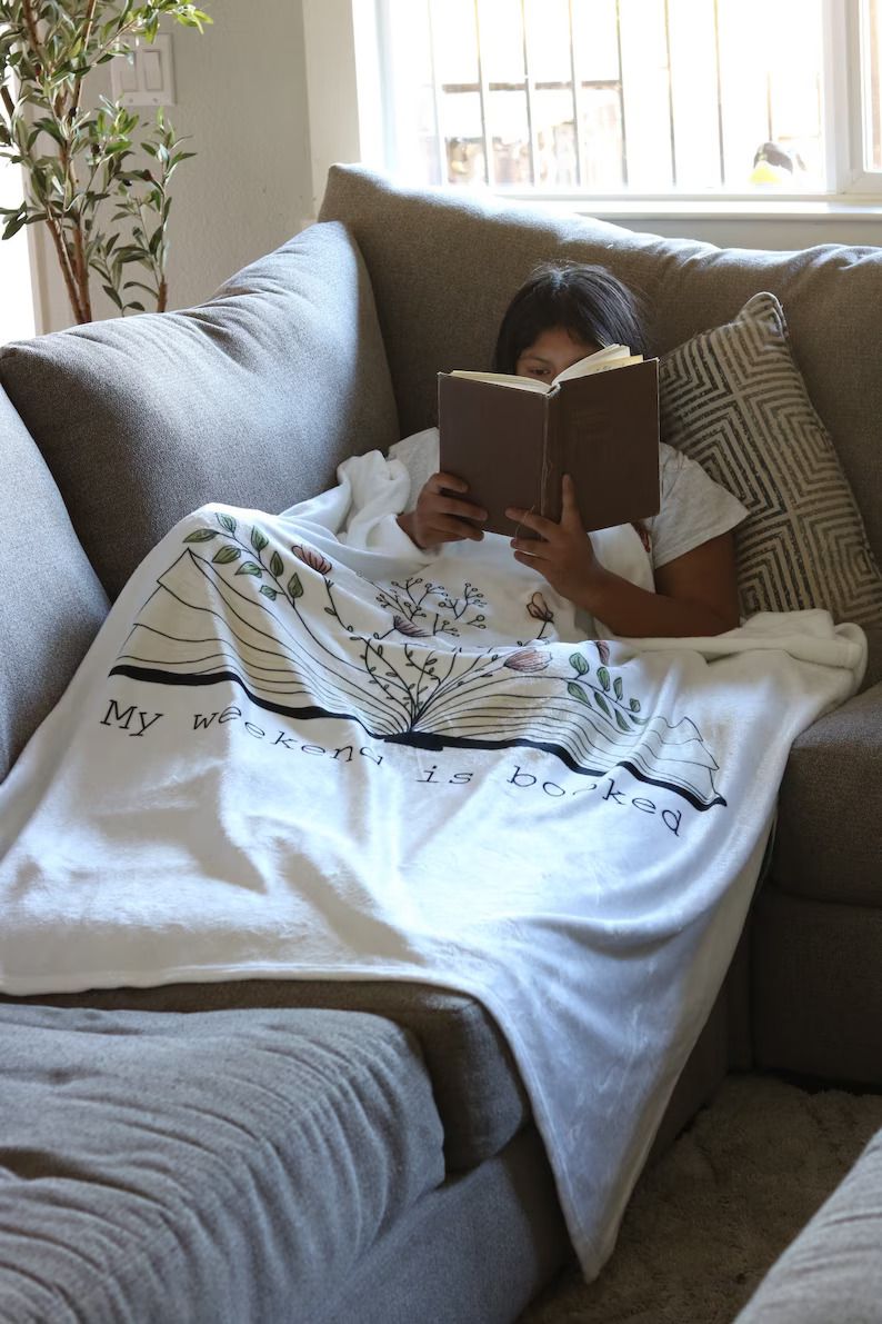 Photo of a person reading a book while laying on the couch, with a blanket on top of them. The blanket has an illustration of an open book from which flowers sprout, and the text My weekend is booked
