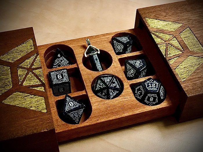 Image of dual opening wooden dice storage box with gold stenciled image on top, holding 8-piece obsidian dice set with white chip glyphic