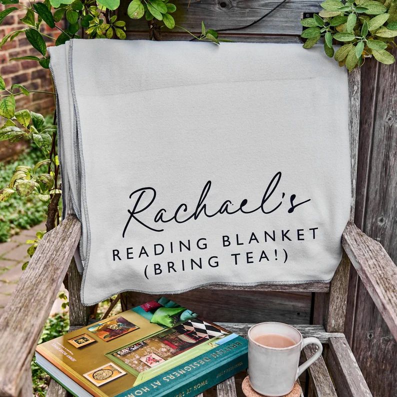 Photo of a blanket folded over a garden chair. The blanket is grey and shows the text Rachael's reading blanket (bring tea!)