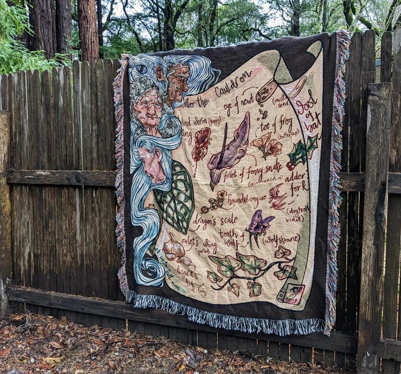 Photo of a blanket placed on a fence showing a gorgeous illustration of the Witches by Macbeth with some text and nature elements