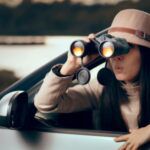 woman with fair skin leaning outside of a car looking curiously through binoculars