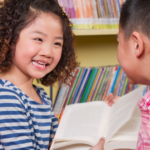 a photo of two Asian children smiling and looking at a book together at a library